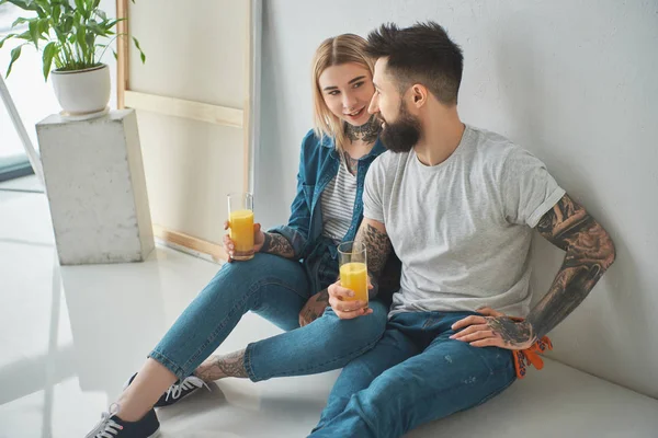 Feliz joven pareja sosteniendo vasos de jugo y sentado en el suelo en nuevo hogar - foto de stock