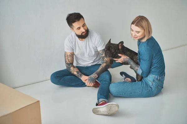 Vue grand angle de sourire jeune couple tatoué assis sur le sol et jouer avec chien dans une nouvelle maison — Photo de stock