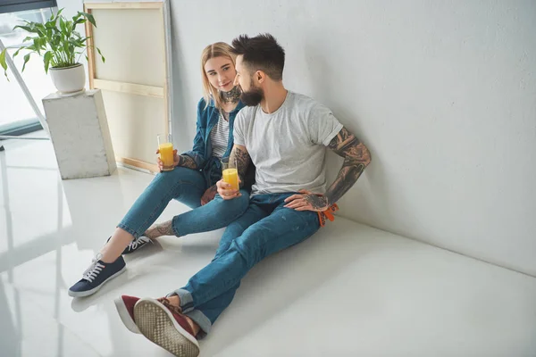 Heureux jeune couple tatoué tenant des verres de jus et assis sur le sol dans une nouvelle maison — Photo de stock