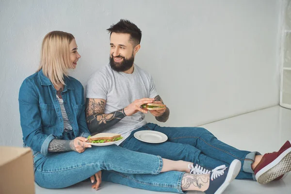 Feliz jovem casal segurando sanduíches e sorrindo uns aos outros na nova casa — Fotografia de Stock