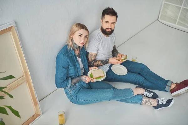 Vue grand angle de jeune couple heureux tenant des sandwichs et souriant à la caméra dans une nouvelle maison — Photo de stock
