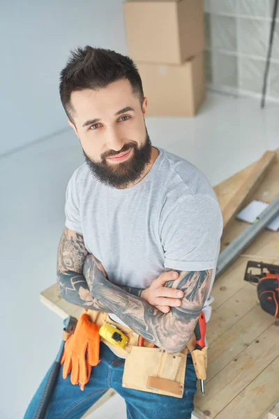 Reparador sonriente con tatuajes y herramientas apoyadas en la superficie de madera en el nuevo apartamento - foto de stock