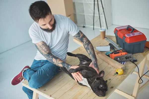 Tätowierter Mann spielt in neuer Wohnung mit französischer Bulldogge auf Holzoberfläche — Stockfoto