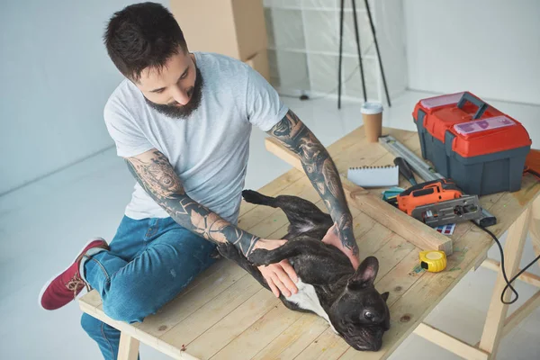 Hombre tatuado jugando con bulldog francés en superficie de madera en nuevo apartamento - foto de stock