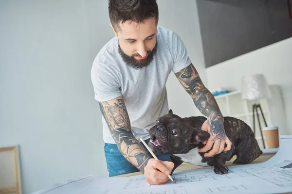 Reparador tatuado com lápis e bulldog francês olhando para o modelo de novo apartamento em mesa de madeira — Fotografia de Stock