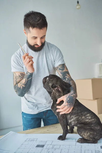 Tattooed repairman with pencil and french bulldog standing at blueprint of new apartment on wooden tabletop — Stock Photo