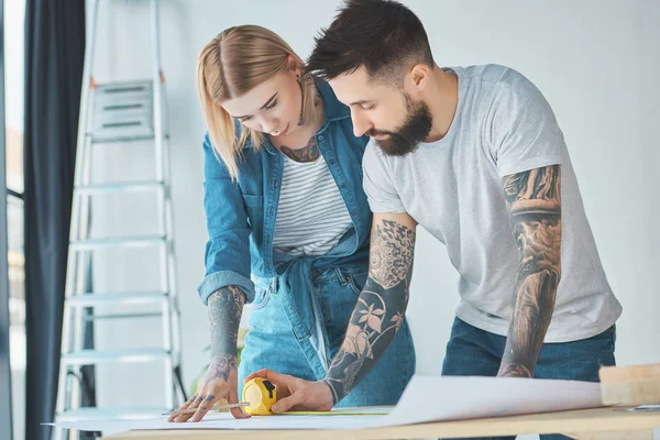 Portrait of tattooed couple looking at blueprints on wooden tabletop at new home — Stock Photo