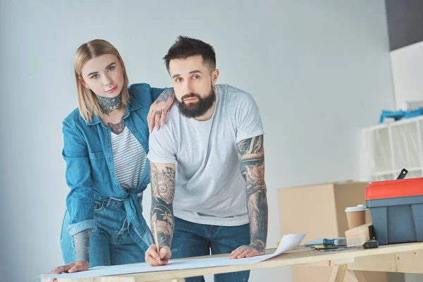 Porträt eines tätowierten Paares, das in die Kamera blickt, während es im neuen Zuhause an Bauplänen auf einer hölzernen Tischplatte steht — Stockfoto
