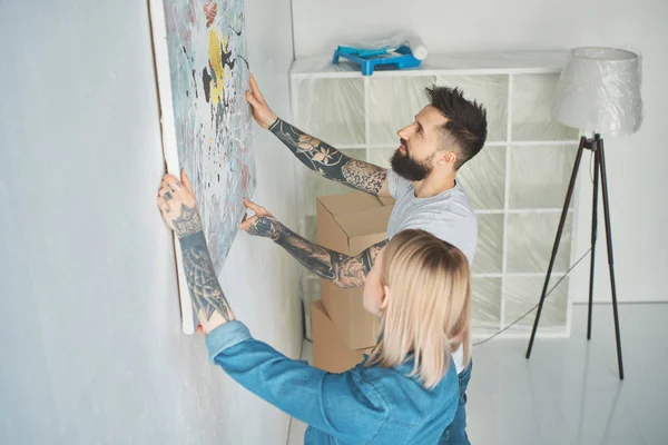 Seitenansicht eines jungen tätowierten Paares, das in seiner neuen Wohnung Gemälde an die Wand hängt — Stockfoto