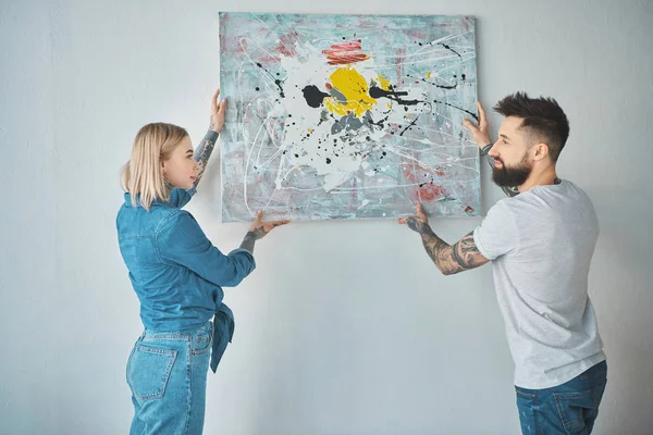 Young tattooed couple hanging painting on wall together at new apartment — Stock Photo