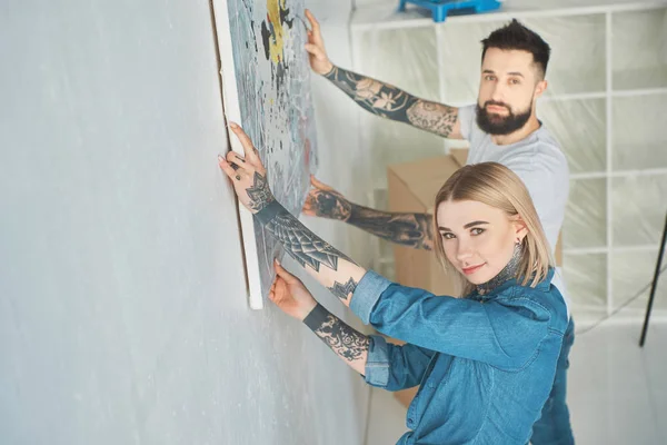 Joven pareja tatuada colgando pintura en la pared en nuevo apartamento - foto de stock