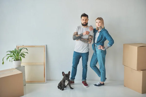 Young couple standing at new home with cardboard boxes and french bulldog — Stock Photo