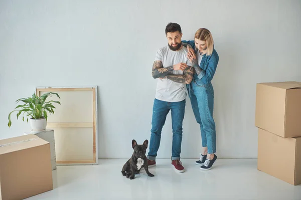 Young couple standing at new home with cardboard boxes and french bulldog — Stock Photo