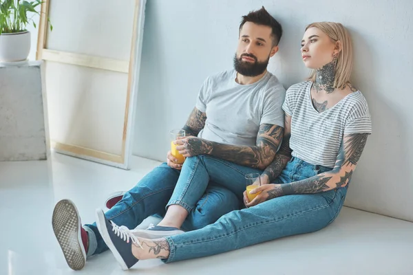 Müdes tätowiertes Paar mit Gläsern Saft sitzt im neuen Zuhause auf dem Boden — Stockfoto