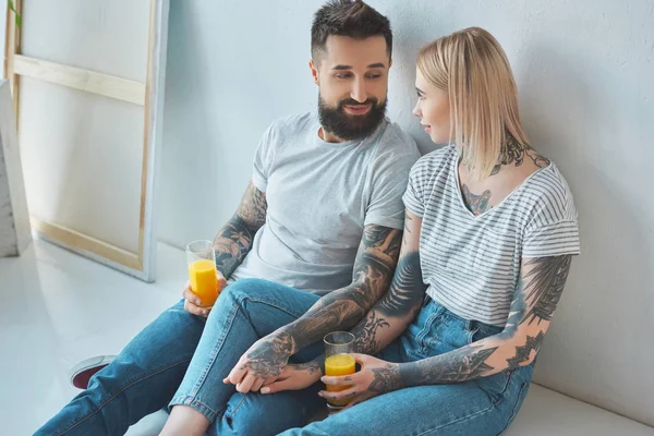 Tattooed couple with glasses of juice holding hands while sitting on floor at new home — Stock Photo