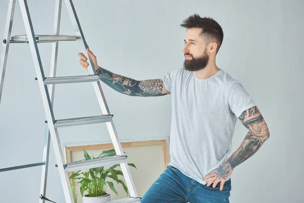 Giovane uomo barbuto con tatuaggi in piedi alla scala nella nuova casa — Foto stock