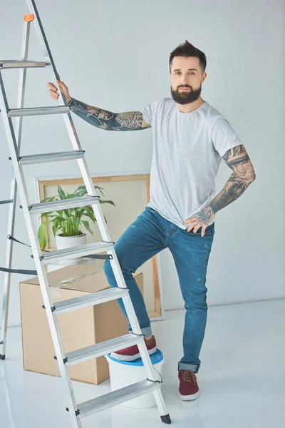 Joven barbudo hombre con tatuajes de pie en la escalera en nuevo hogar - foto de stock