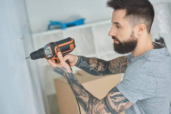 Tatuado homem fazendo buraco na parede com parafuso arma no novo apartamento, inexperiente conceito milenar — Fotografia de Stock