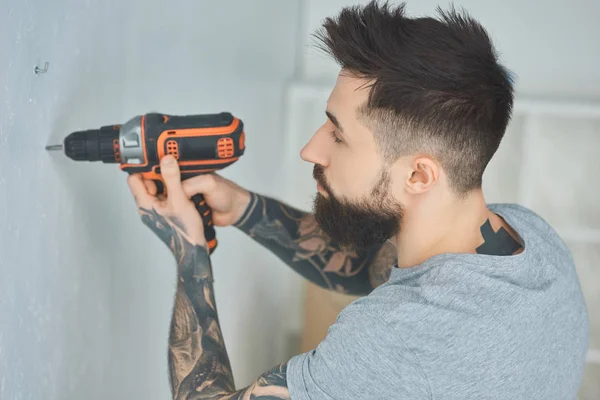 Татуированный человек делает дыру в стене с винтовым пистолетом в новой квартире, неопытная концепция тысячелетия — стоковое фото