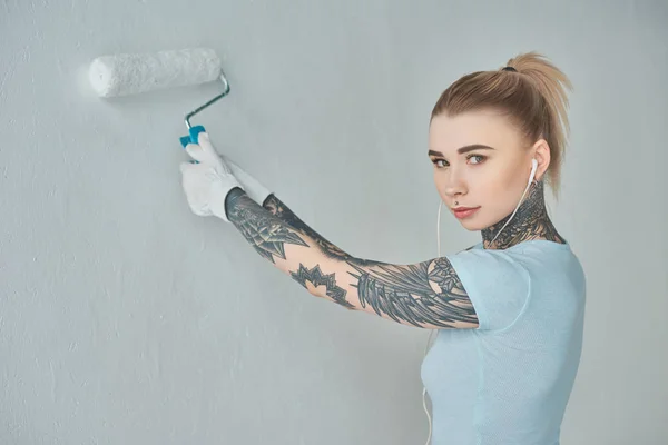 Mujer joven tatuada en auriculares pintando la pared en un nuevo hogar - foto de stock