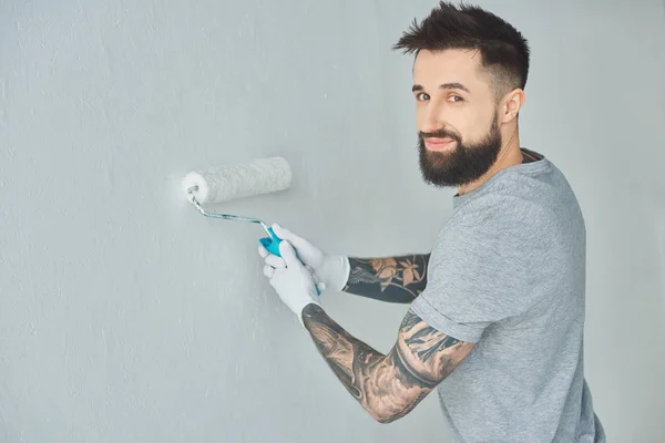 Sonriente reparador tatuado con pincel de pintura en rollo mirando a la cámara mientras pinta la pared en un apartamento nuevo - foto de stock