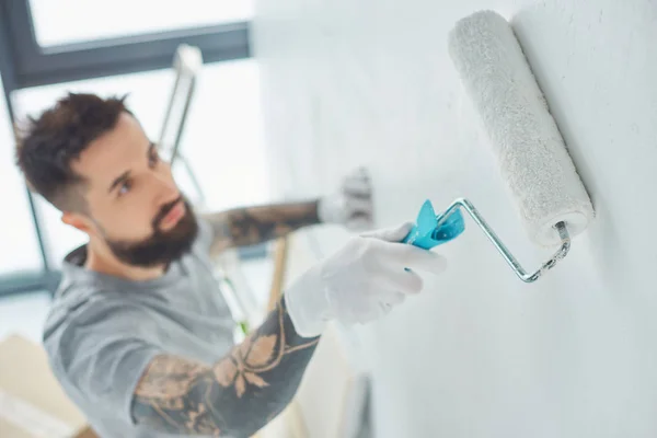 Selective focus of tattooed repairman with roll paint brush painting wall in new apartment — Stock Photo