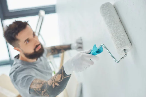 Enfoque selectivo de reparador tatuado con pared de pintura de pincel de rollo en nuevo apartamento - foto de stock