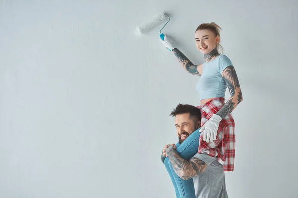 Smiling woman with roll paint brush sitting on boyfriends shoulders while painting wall at new home — Stock Photo