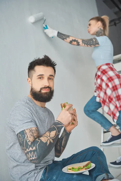 Tätowierter Mann isst Sandwich, während Freundin Wand im neuen Zuhause bemalt — Stockfoto