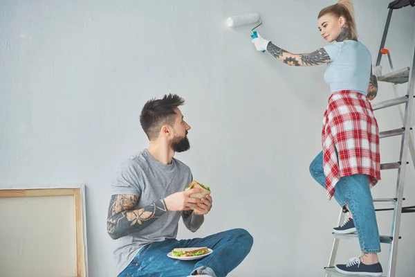 Татуйований чоловік їсть бутерброд, а дівчина на драбині малює стіну в новому будинку — стокове фото