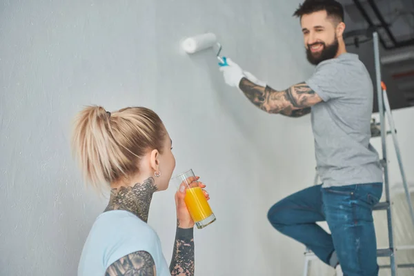 Joven mujer beber jugo mientras novio en escalera pintura pared en nuevo apartamento - foto de stock