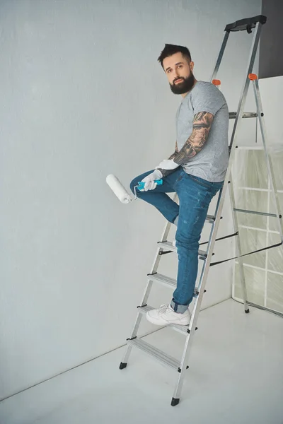 Jeune réparateur tatoué avec pinceau rouleau debout sur l'échelle dans un nouvel appartement — Photo de stock