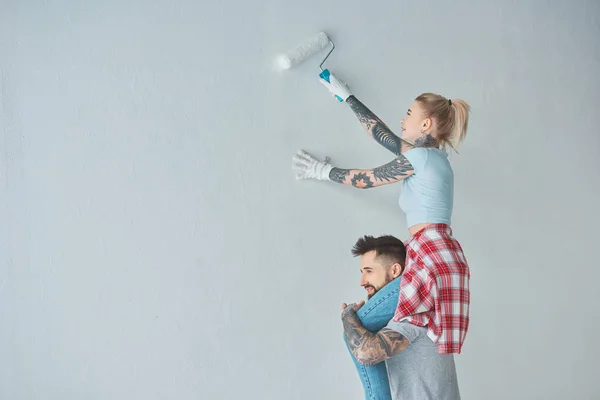 Woman with roll paint brush sitting on boyfriends shoulders while painting wall at new home — Stock Photo