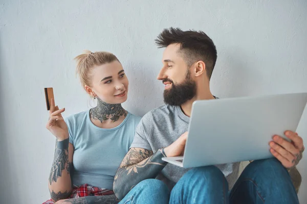 Retrato de pareja tatuada con ordenador portátil y tarjeta de crédito compras en línea en el nuevo apartamento - foto de stock