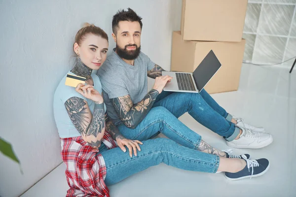 Junges Paar mit Laptop und Kreditkarte blickt beim Online-Shopping in neue Wohnung in die Kamera — Stockfoto