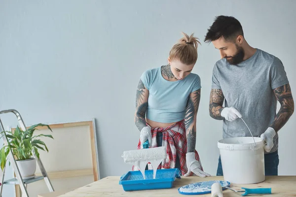 Pareja tatuada con pinceles de pintura en rollo haciendo reparaciones en el nuevo apartamento - foto de stock