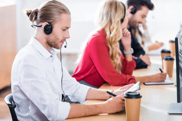 Foco seletivo dos operadores de call center em headsets com notebooks trabalhando no local de trabalho no escritório — Fotografia de Stock