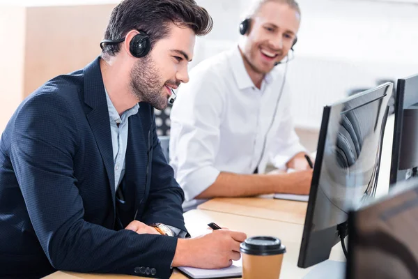 Enfoque selectivo de los operadores de centros de llamadas masculinos en auriculares con cuadernos en el lugar de trabajo en la oficina - foto de stock