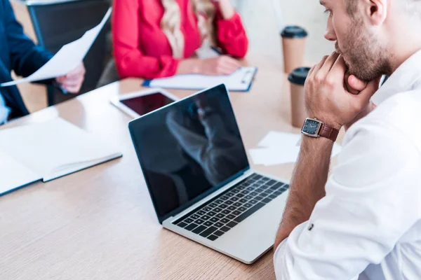 Partial view of pensive businessman looking at blank laptop screen at workplace with papers and colleagues near by — Stock Photo