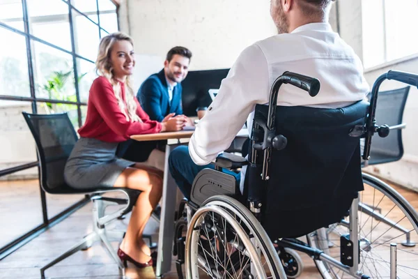 Частичный взгляд улыбающихся бизнесменов на коллегу в инвалидной коляске в офисе — стоковое фото