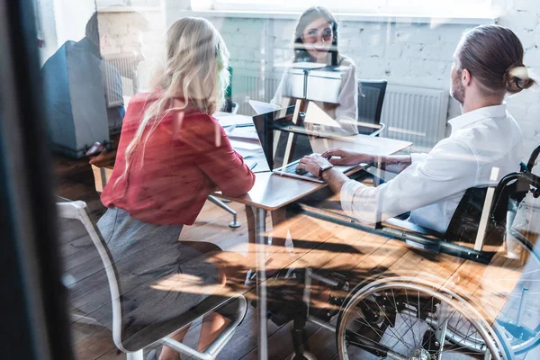 Vue à travers le verre de jeune homme d'affaires handicapé en fauteuil roulant travaillant avec des collègues au bureau — Photo de stock