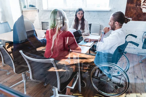 Vista através de vidro de jovem empresário em cadeira de rodas que trabalha com colegas no escritório — Fotografia de Stock