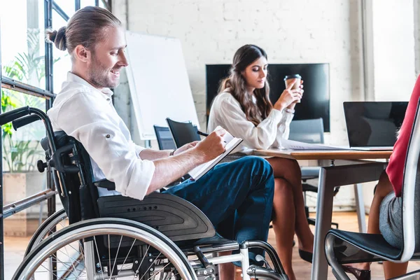 Улыбающийся бизнесмен-инвалид в инвалидной коляске делает заметки на рабочем месте — Stock Photo