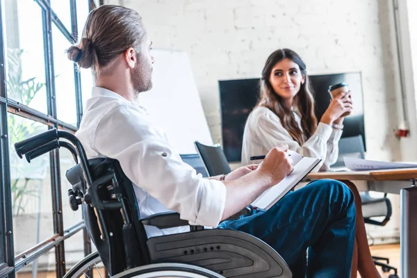 Young businessman in wheelchair writing in notebook and working with smiling colleague in office — Stock Photo