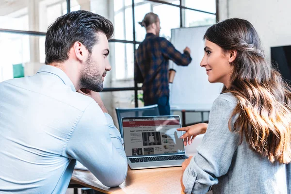 Jóvenes empresarios que utilizan el ordenador portátil con el sitio web de noticias de la BBC en la pantalla y sonriendo entre sí en el lugar de trabajo - foto de stock