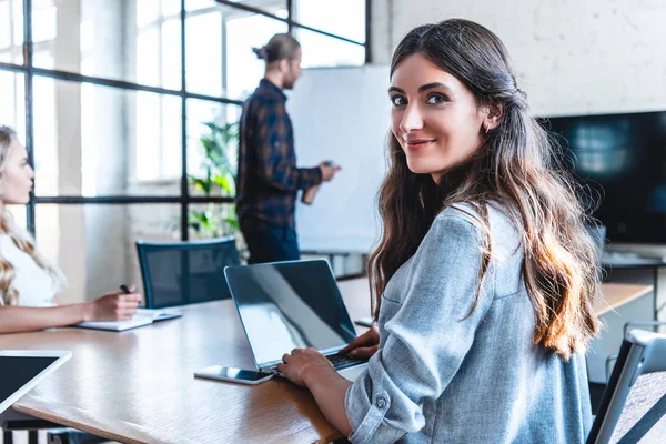 Young businesswoman using laptop and smiling at camera while working with colleagues in office — Stock Photo