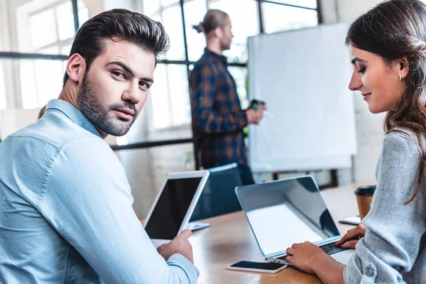 Jovens empresários usando dispositivos digitais, enquanto colega do sexo masculino escrevendo no quadro branco atrás — Fotografia de Stock