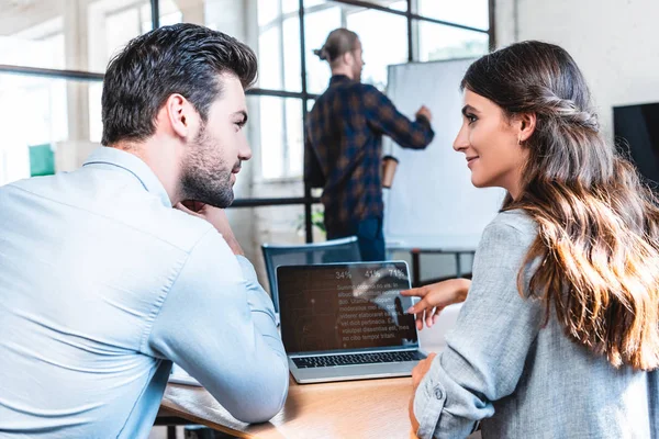 Junge Geschäftsleute, die mit Laptop arbeiten und einander anschauen, während männliche Kollegen hinter Whiteboard schreiben — Stockfoto