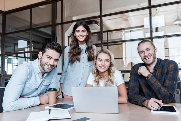 Jóvenes colegas de negocios felices trabajando con gadgets y sonriendo a la cámara en la oficina - foto de stock