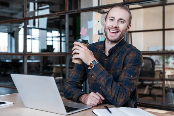 Guapo joven empresario sosteniendo taza de papel y sonriendo a la cámara mientras trabaja con el ordenador portátil en la oficina - foto de stock